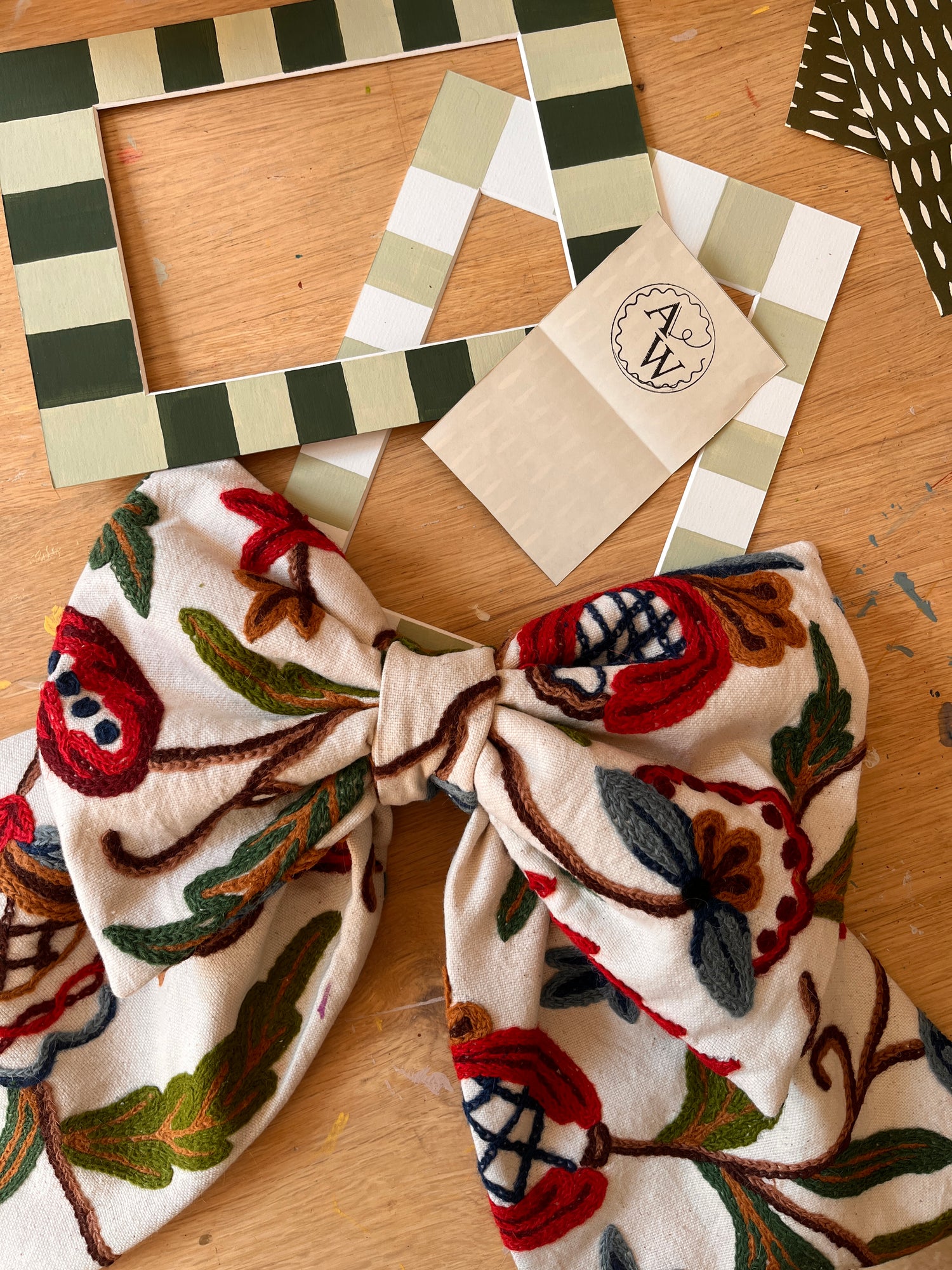 Decorative striped picture mount mat next to a Annabel Wond Interiors branded card and a handmade embroidered fabric bow fabric with floral patterns on a wooden surface