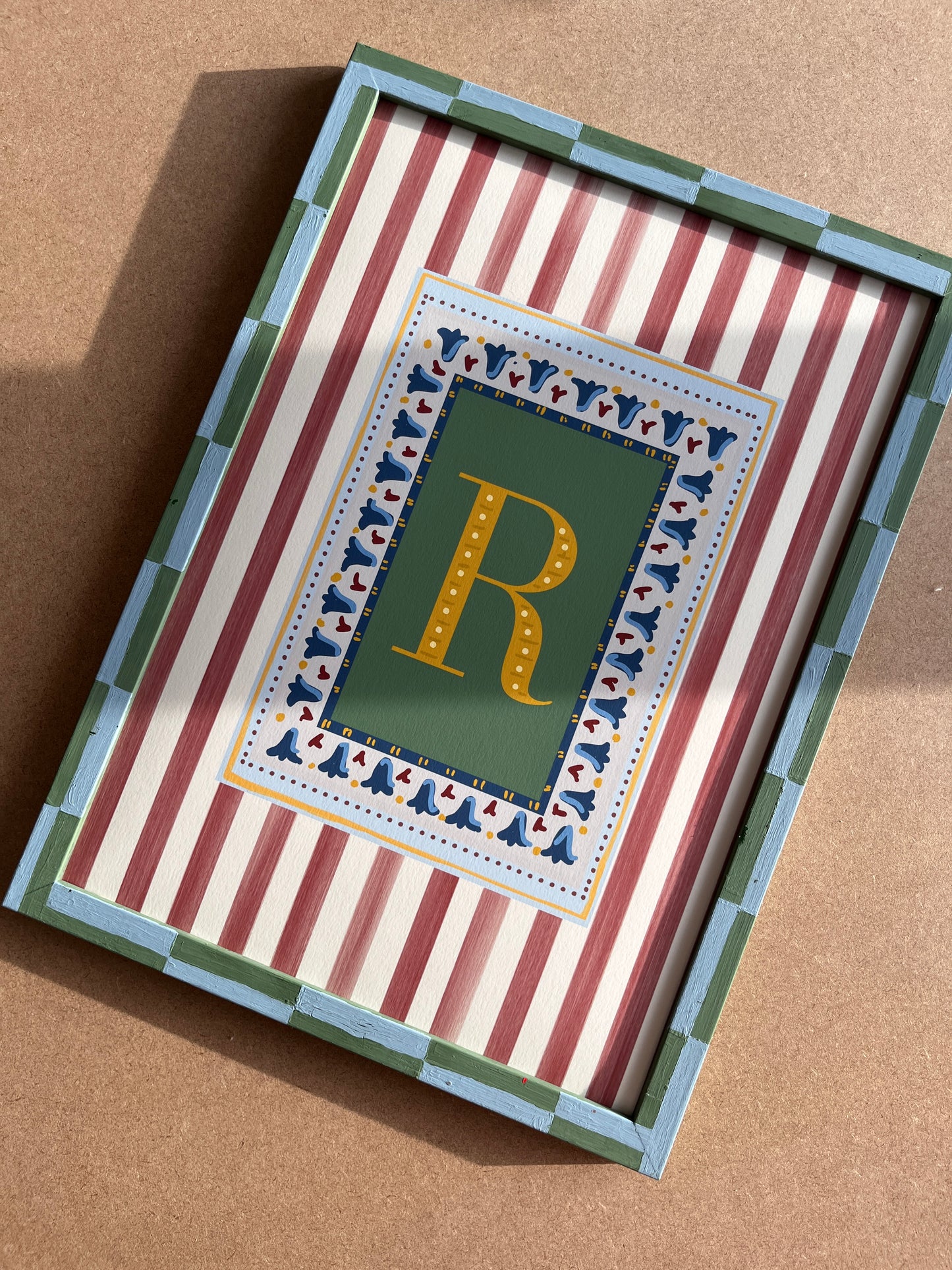 Nursery alphabet wall art featuring the letter of your choice a golden serif font  with green backdrop and decorative block-print inspired folk floral border, framed by a muted red striped background and finished with a hand painted checkered frame.  Perfect for baby room decor, personalized children’s wall art, alphabet nursery print, gender-neutral baby gift, Montessori room decor, educational wall art, and unique custom gift ideas for kids and adults. 
