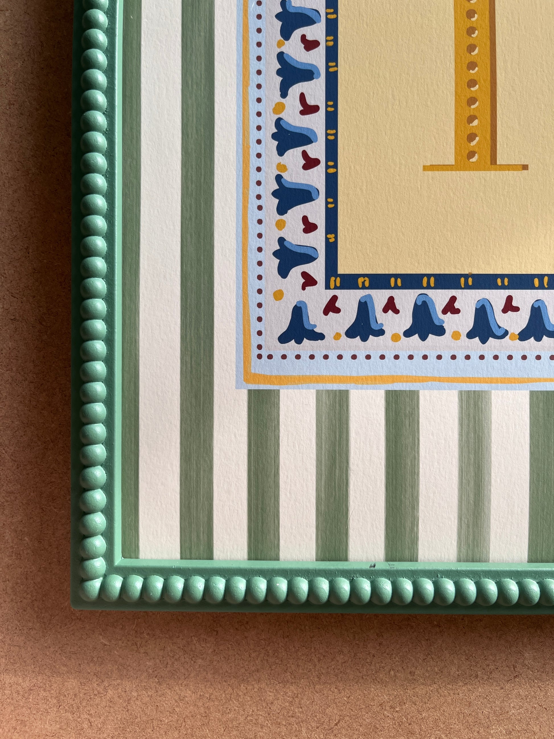 Nursery alphabet wall art featuring the letter of your choice a golden serif font with decorative block-print inspired folk floral border and a muted green striped background, perfect for baby room decor, personalized children’s wall art, alphabet nursery print, gender-neutral baby gift, Montessori room decor, educational wall art, and unique custom gift ideas for kids