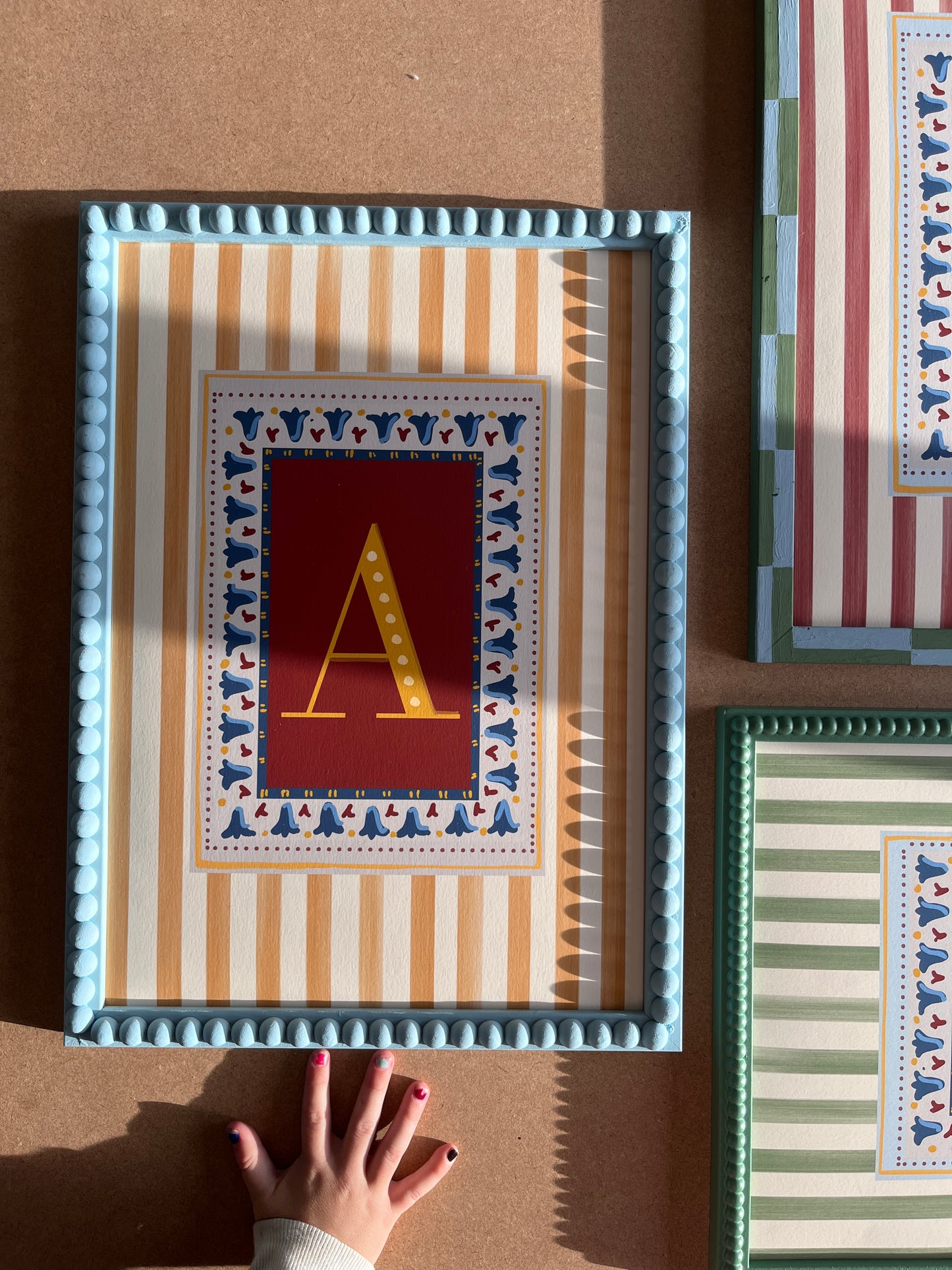 Decorative and unique alphabet wall art featuring your chosen letter in a bold yellow font on a deep red background, framed with a floral block-print style border and set against vertical muted yellow stripes—ideal for playful nursery decor, alphabet print collections, and unique gift ideas for children’s playrooms and nursery decor as well as design lovers