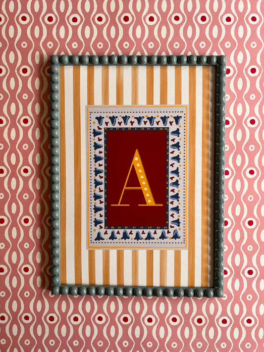 A decorative letter art print with the letter in yellow on a rust background surrounded by a yellow striped border, framed with a blue bobbin frame with a colourful pink patterned backdrop. 