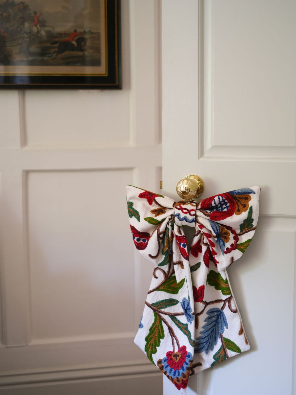 Hand-embroidered vintage fabric crewel work bow with vibrant floral and vine motifs in red, green, blue, and ochre, styled in a traditional countryside cotswold home — perfect as a decorative accent for furniture,  door knobs, the garden or seasonal home styling, it would also make a unique gift 
