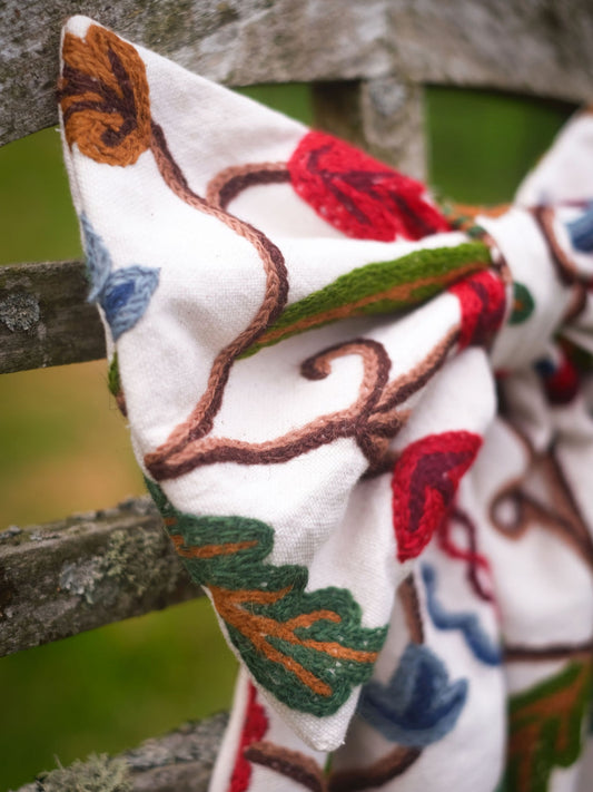 Hand-embroidered vintage fabric crewel work bow with vibrant floral and vine motifs in red, green, blue, and ochre, styled in a traditional countryside cotswold home garden— perfect as a decorative accent for furniture,  door knobs, the garden or seasonal home styling, it would also make a unique gift 