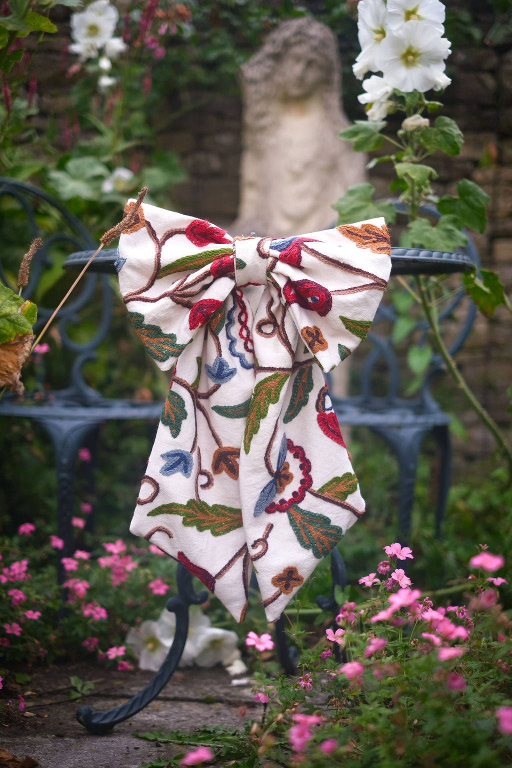 Hand-embroidered vintage fabric crewel work bow with vibrant floral and vine motifs in red, green, blue, and ochre, styled on a vintage wrought iron chair in a garden setting — perfect as a decorative accent for furniture,  the garden or seasonal home styling, it would also make a unique gift 