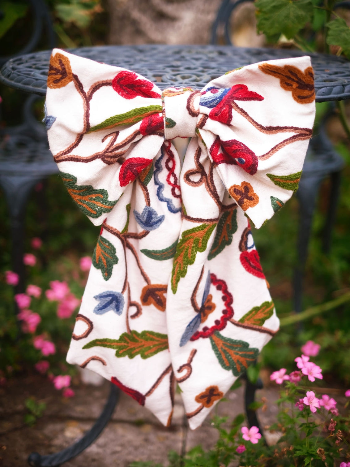 Hand-embroidered vintage fabric crewel work bow with vibrant floral and vine motifs in red, green, blue, and ochre, styled in a traditional countryside cotswold home — perfect as a decorative accent for furniture,  door knobs, the garden or seasonal home styling, it would also make a unique gift 