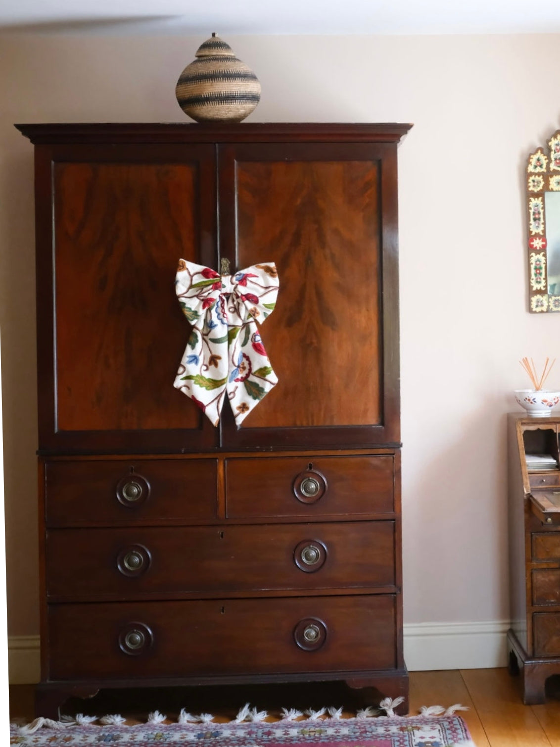 Floral handmade decorative fabric bow made from embroidered crewel work tied to the front of a vintage wooden armoire, adding a soft, handcrafted touch and seasonal charm to classic furniture styling — ideal for home decor accents, festive dressing, or unique interior personalization.