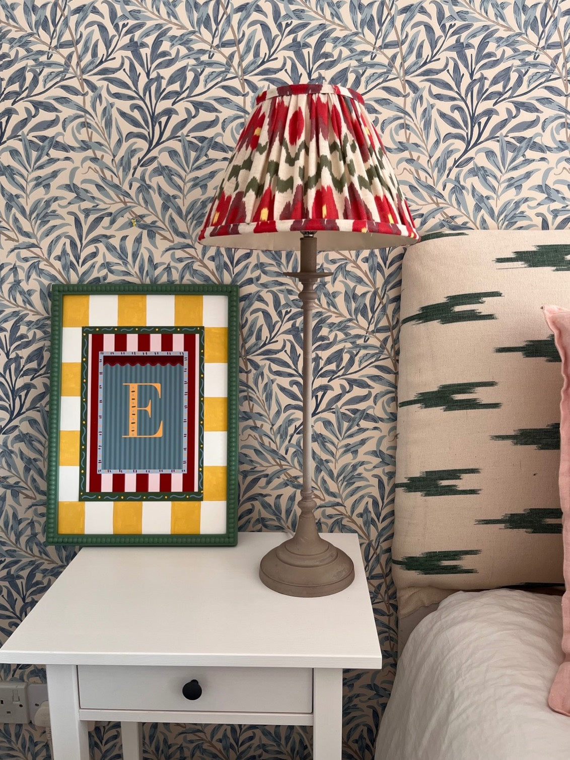 Framed alphabet wall art featuring the letter ‘B’ in yellow on a blue striped background with colorful decorative borders. Paired here with a yellow and white striped, hand painted picture mount/mat and green bobbin frame perfect for nursery decor, kids and baby room styling, or unique alphabet print gift idea. Decorative interiors backdrop and ikat lampshade and headboard. 
