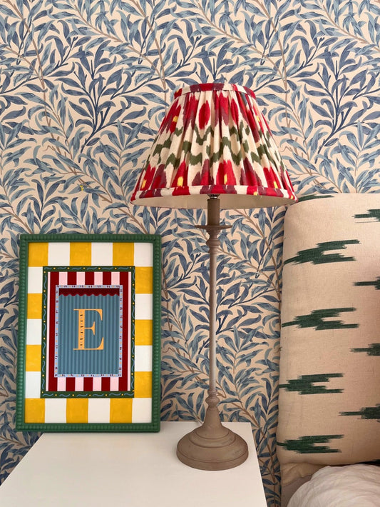 Decorative alphabet print with stripe mount and hand painted bobbin frame next to an ikat  lamp with a colourful shade on a bedside table against a patterned wall.