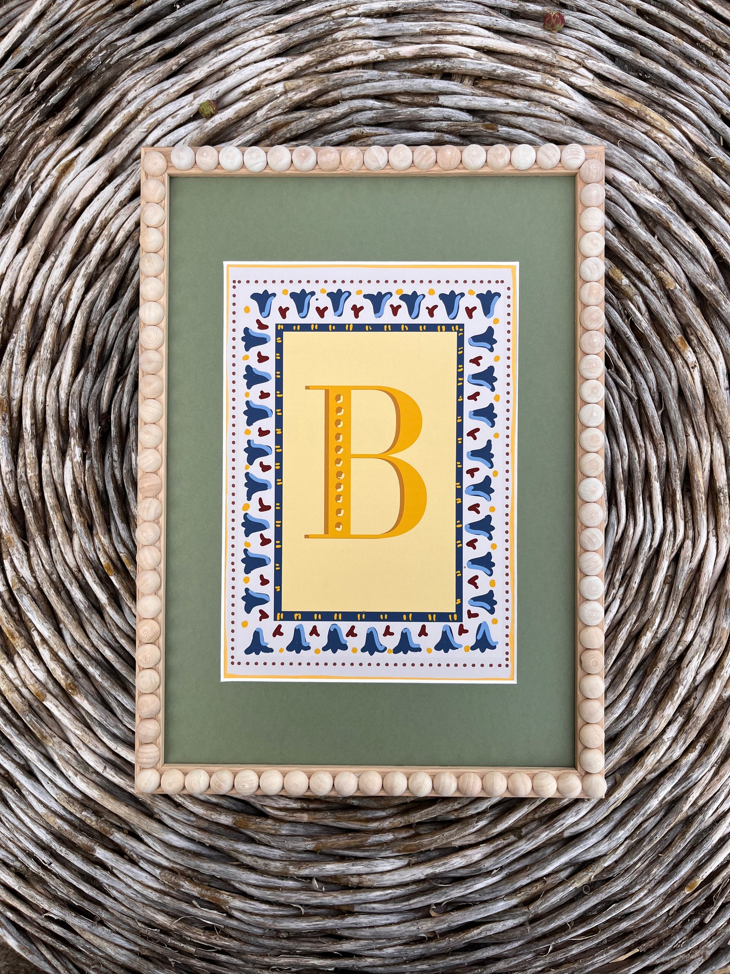 Personalised vintage initial letter print featuring a mustard yellow alphabet letter on a muted sage green background with blue floral border and dusty red striped detailing, styled in a light wooden bobbin frame for a gender-neutral nursery or children's bedroom.